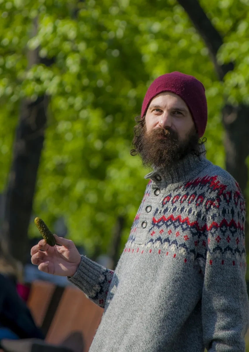 Bearded man in a burgundy knit cap holding a pickle in a sunlit park